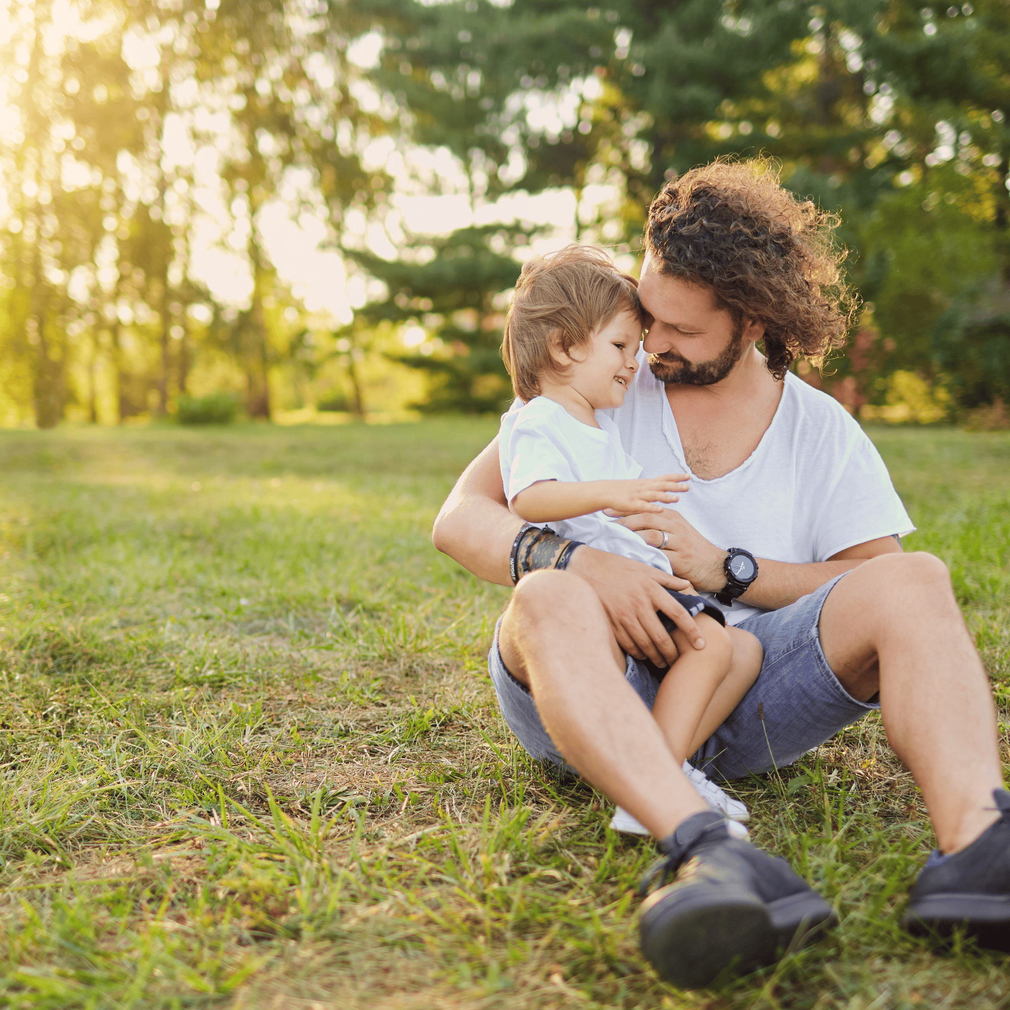 A father lovingly embraces his young son in a sunny park. Sitting on grass, they share a tender moment, smiling warmly. Trees are softly blurred in the background. Navigating Your Child's Autism Diagnosis
