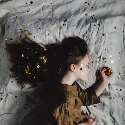 Young girl sleeping peacefully on a white sheet, with dark hair spread around her. Gold star confetti is scattered in her hair and on the sheet. Calm, dreamy mood. Child with a sleep schedule.
