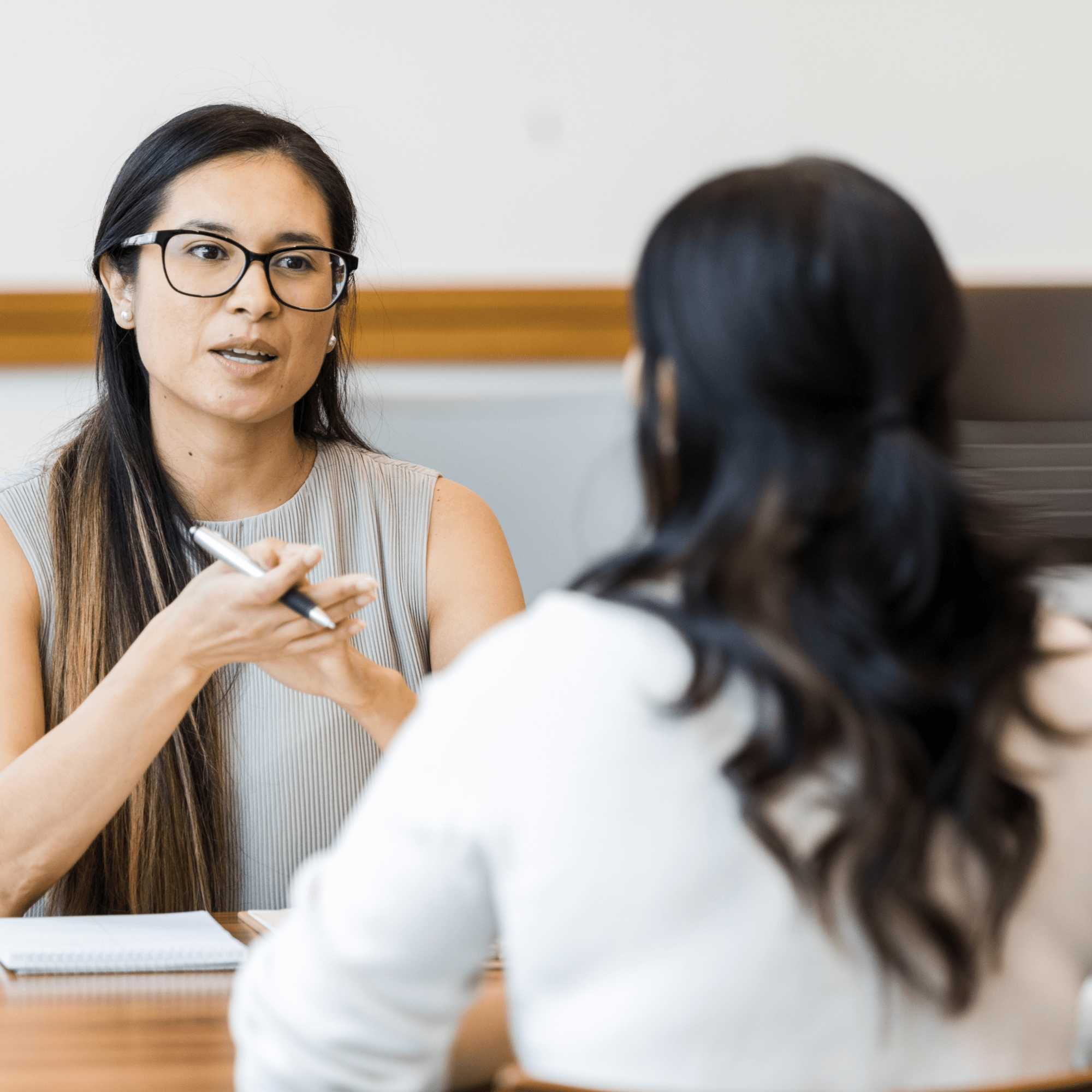 A woman in glasses speaks attentively, as if giving feedback, holding a pen, across from another woman with long hair in an office setting, conveying focus and engagement.