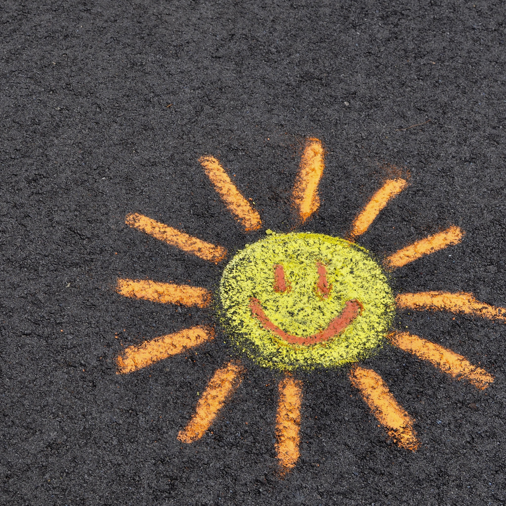 A cheerful sun with a smiling face is drawn in yellow and orange chalk on dark asphalt, conveying a bright and playful tone.