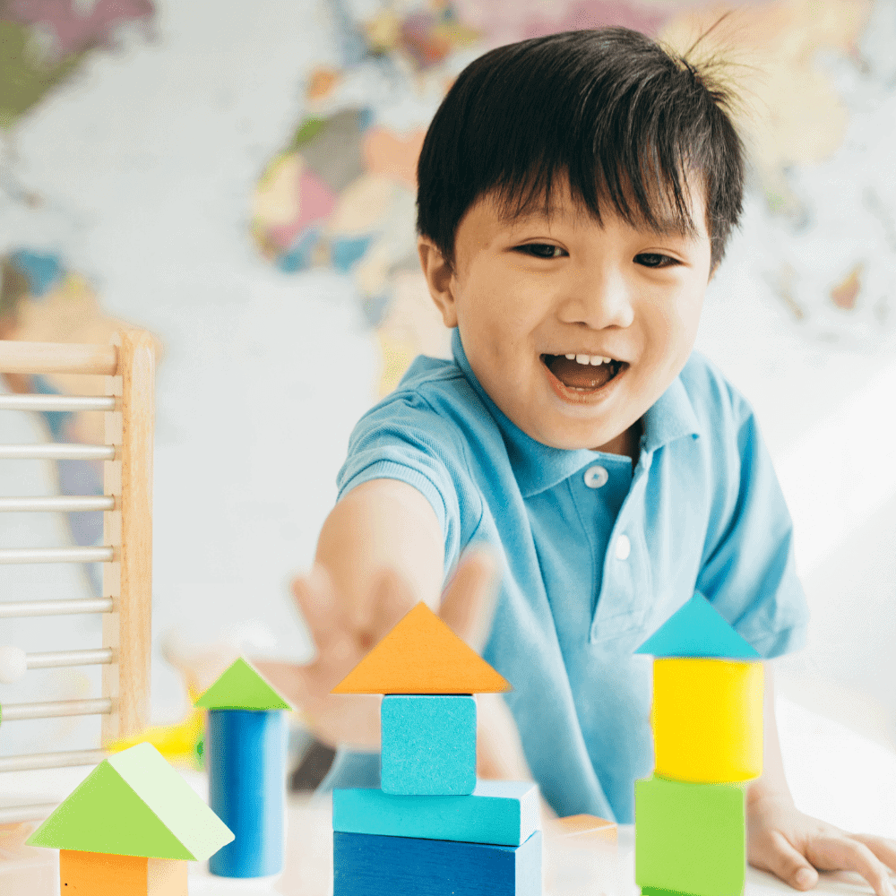A cheerful young boy in a blue shirt engages in early intervention learning activities, playing with colorful building blocks and smiling, supporting developmental skills in a playful educational setting.
