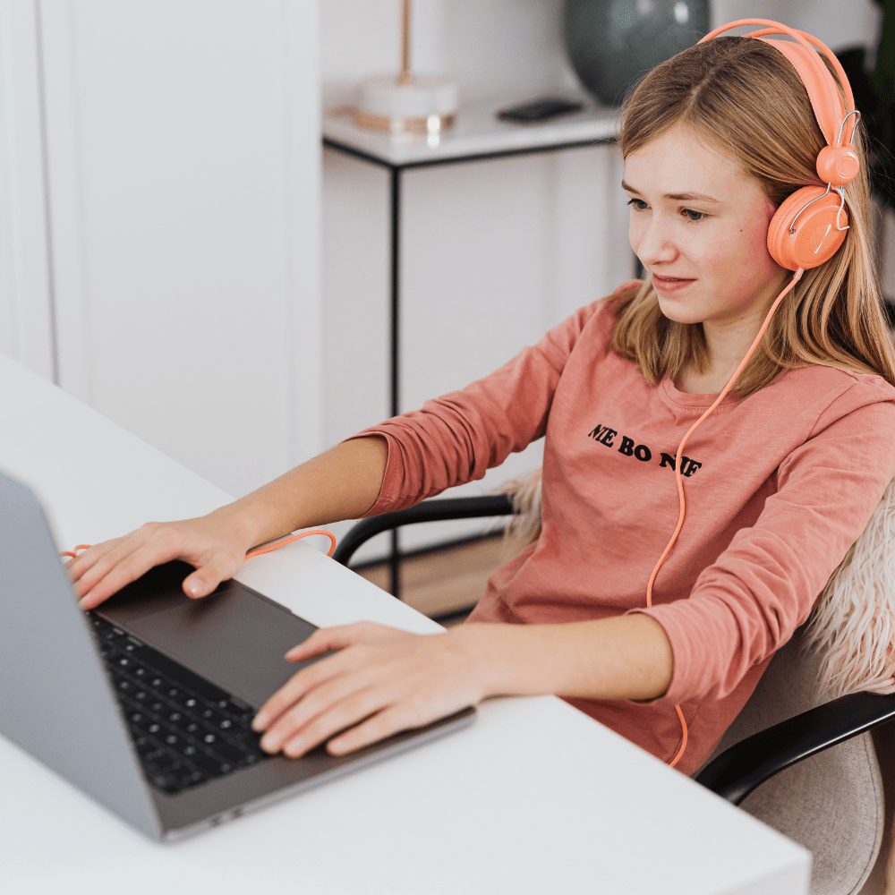 A girl with peach headphones sits at a desk, focusing on a laptop. She's dressed in a pink top in a modern, cozy room, exuding concentration. Sibling Support Group