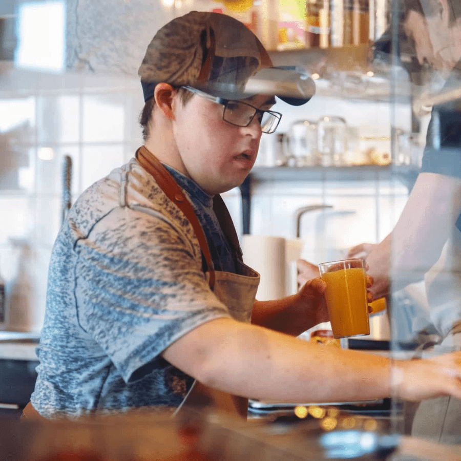 A young man wearing a cap and apron is holding a glass of orange juice in a café. He appears focused, with a busy kitchen in the background.